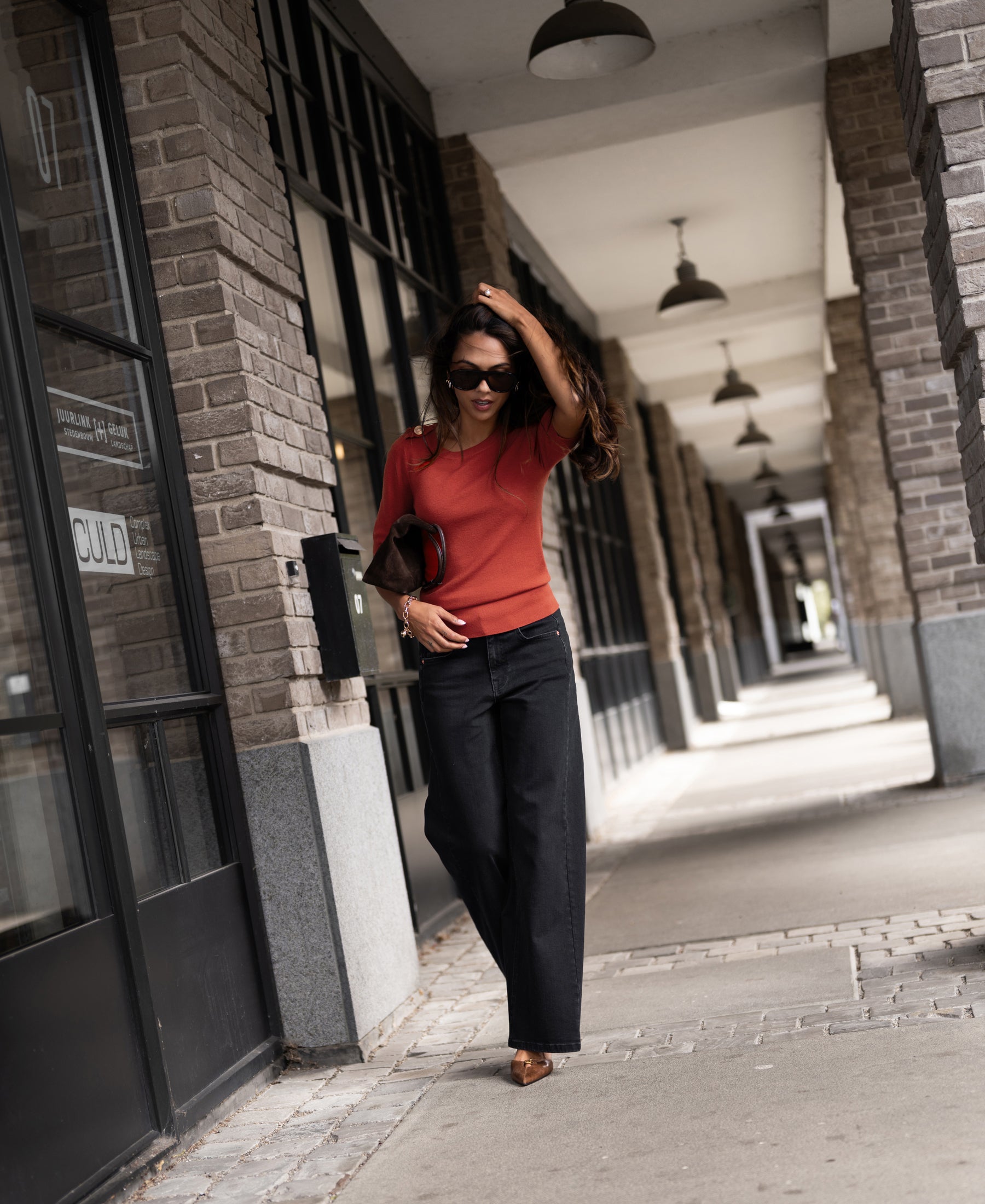 Woman wearing a Merino wool LE MAJOR Chili sweater, shown walking outdoors beside a brick building.