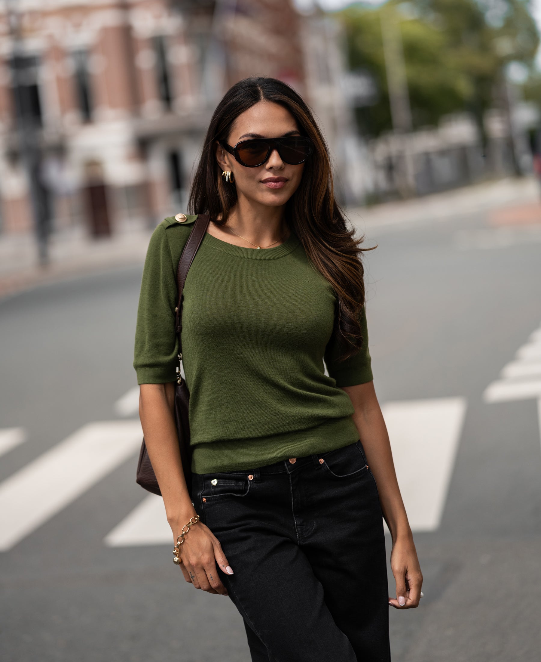 Merino wool sweater in olive green shown on woman outdoors, with trees and buildings visible in the background.