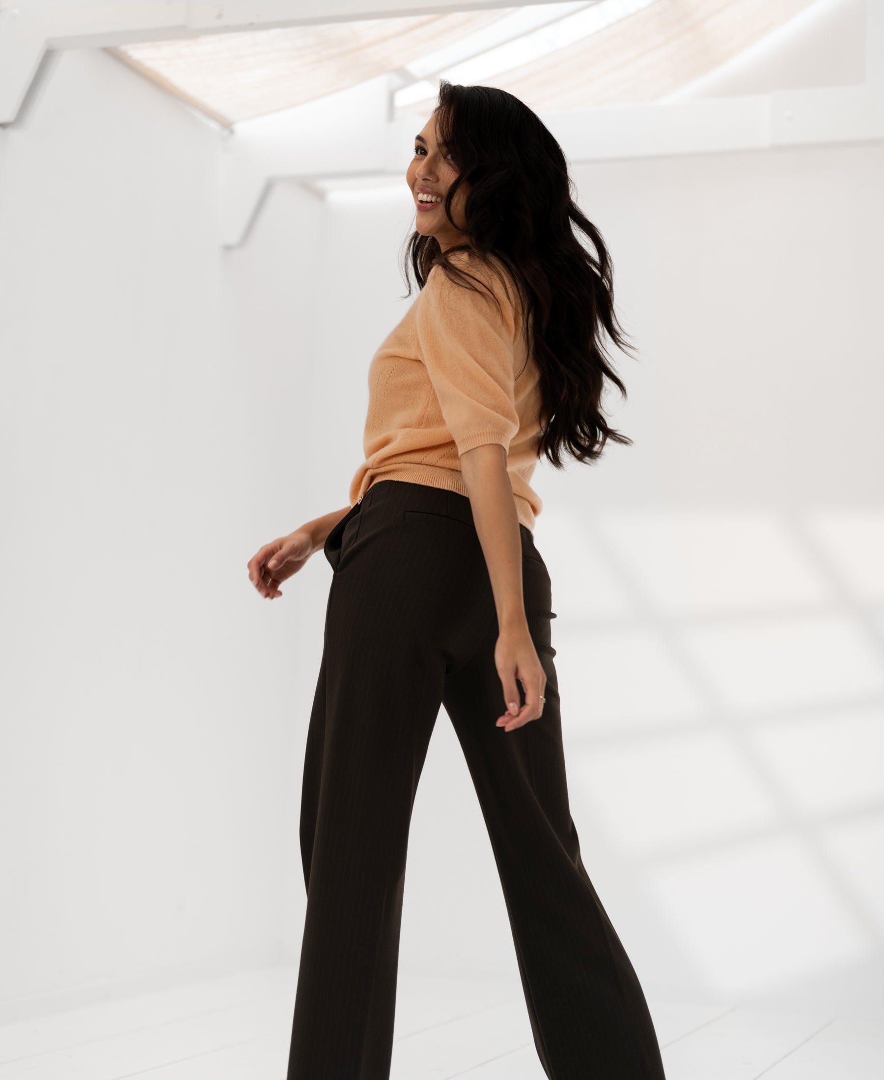 Woman wearing PLEIN PUBLIQUE pinstripe trousers LE MALO in dark brown, standing in a sunlit room.