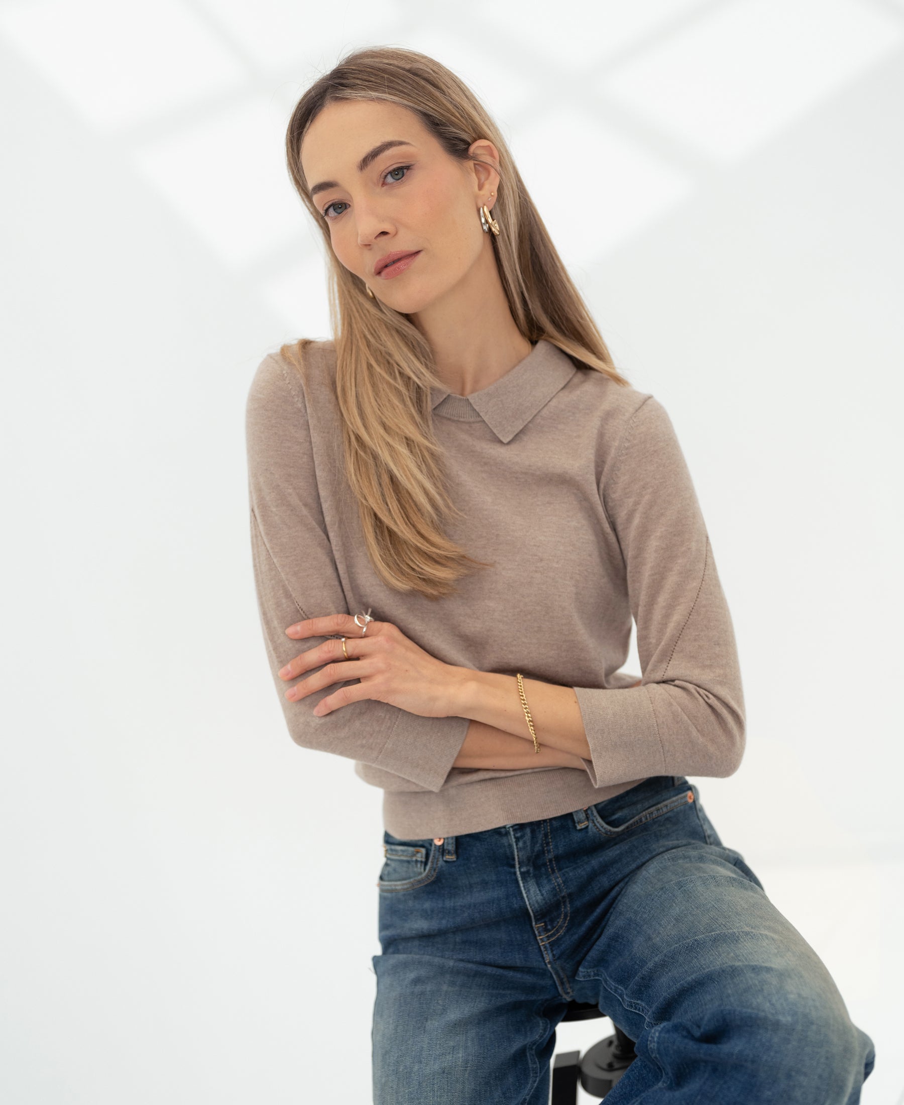 Woman wearing a PLEIN PUBLIQUE merino wool polo collar sweater in sand color, seated.