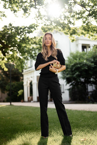 Woman wears black PLEIN PUBLIQUE flared pants standing on grass, with trees and a white house in the background.