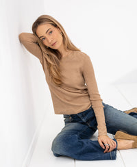 Woman wearing a camel-colored Merino wool shirt sits on the floor, looking at the camera.