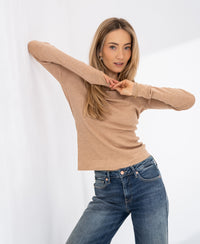Woman wearing a camel merino wool shirt poses with arms raised against a white backdrop.