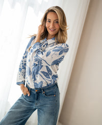 Woman wearing a floral print blouse with tie detail by PLEIN PUBLIQUE, standing by sheer curtains in a bright room.