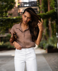 Woman wearing a mocha-colored cupro blouse walks outdoors with greenery in the background.