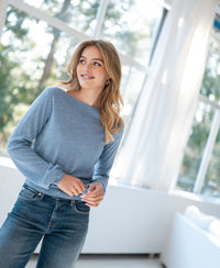 Boat neck LA MUSE Pullover by PLEIN PUBLIQUE in jeans blue, shown indoors with sunlight and windows in the background.