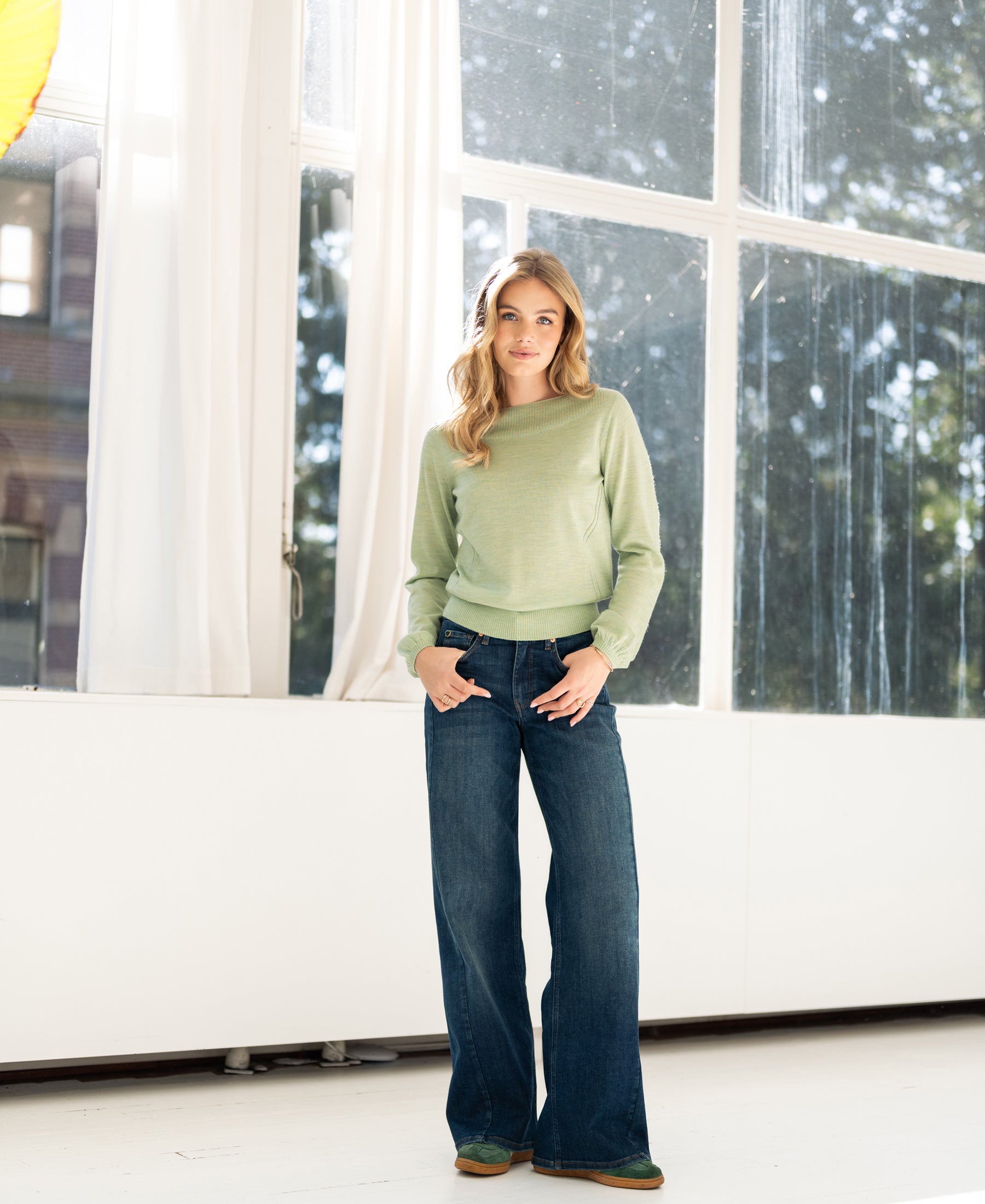 Woman wearing the light green PLEIN PUBLIQUE LA MUSE boat neck sweater, hands in pockets by large sunny windows.