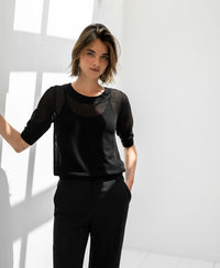 Woman wears Plein Publique LE NANCY mesh silk sweater in black, posing by a sunlit white wall.