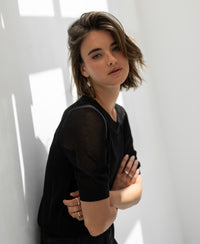 Black mesh sweater with silk, front view. Model stands by a sunlit white wall with arms crossed.
