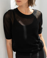 Woman wears black mesh silk sweater by Plein Publique, standing against a white wall.