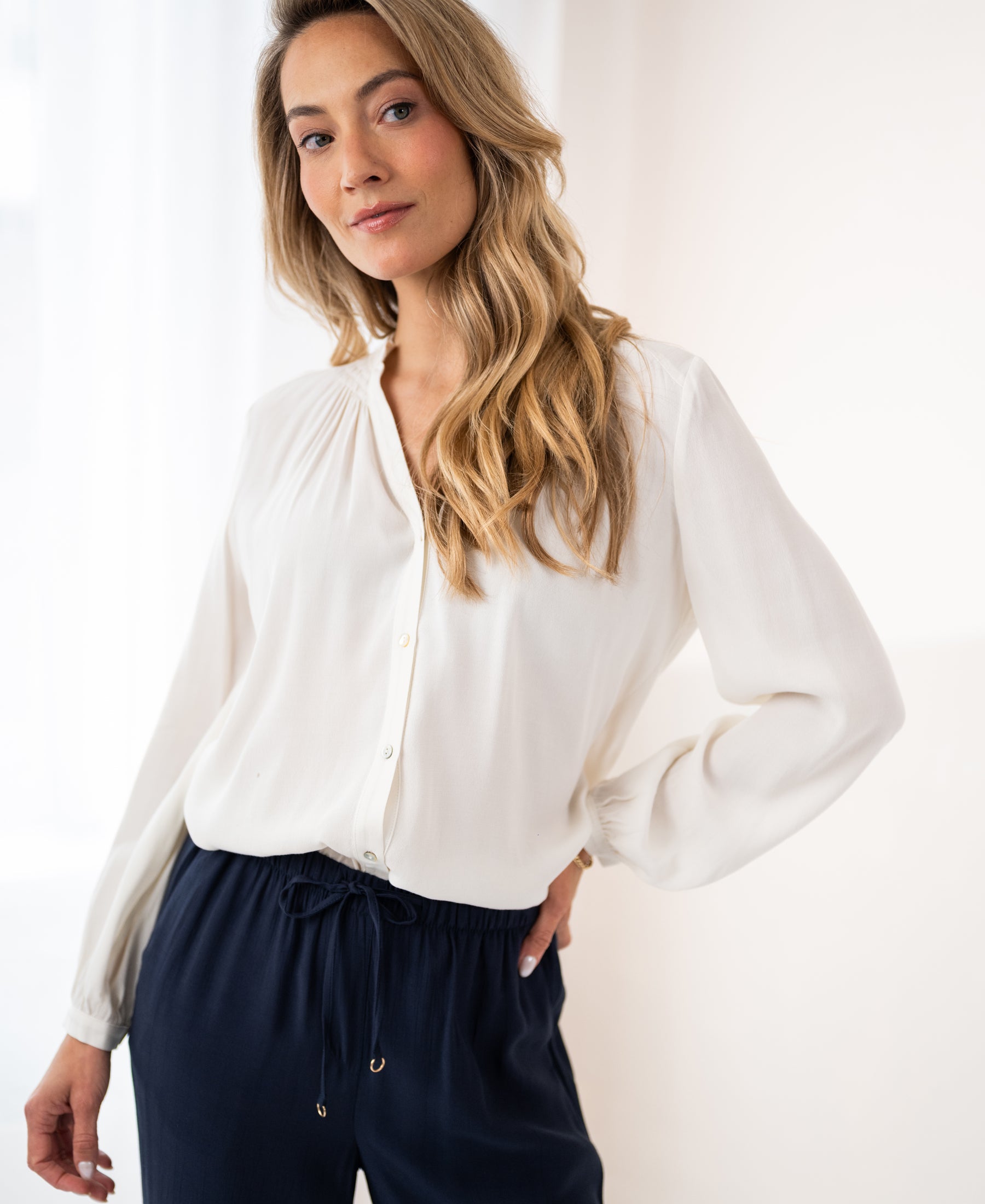 Woman with long blonde hair wears the LA NIMES ivory blouse by PLEIN PUBLIQUE, hand in pocket.