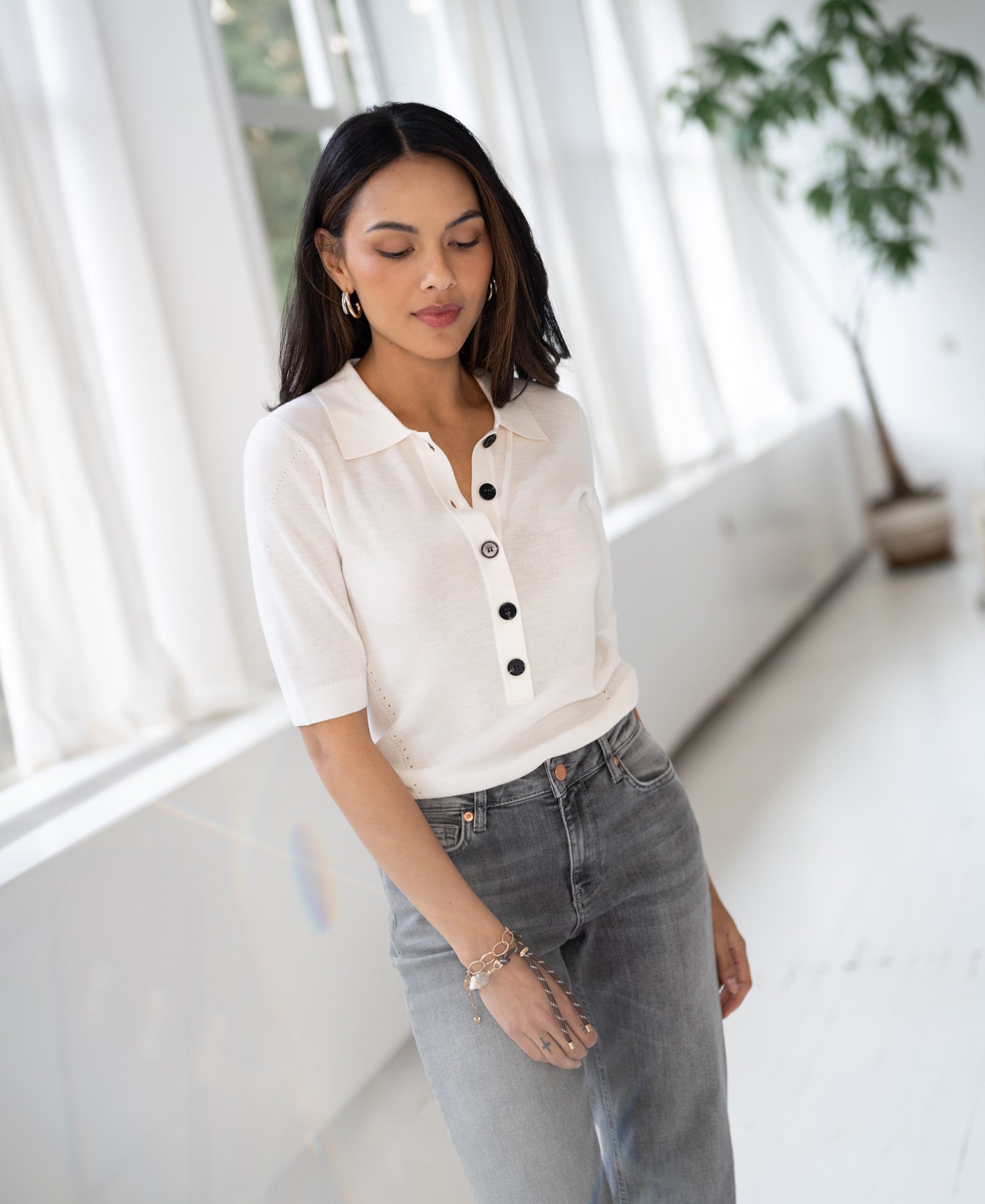 Woman by large windows wears ivory merino wool polo shirt, eyes gently closed.