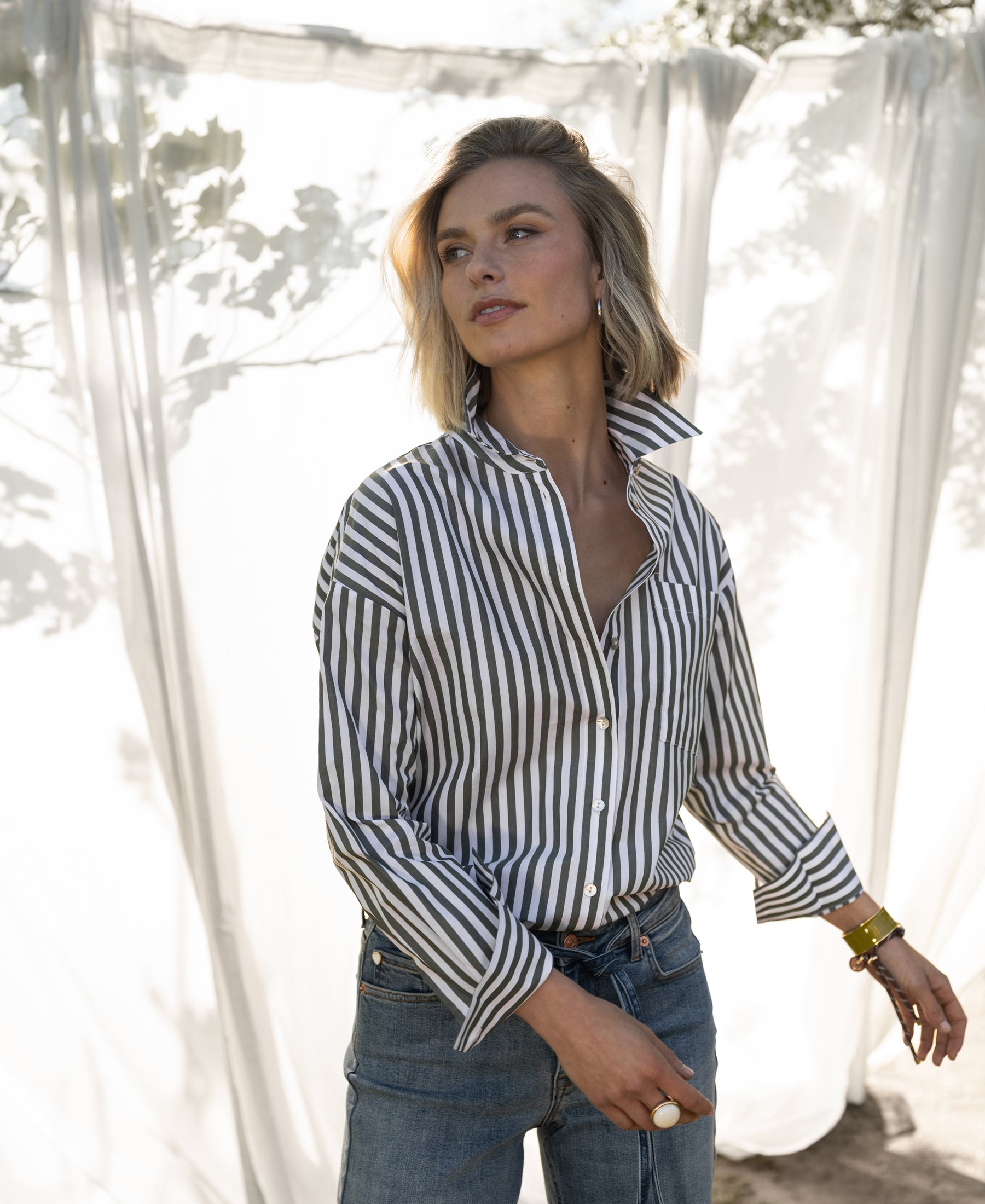 Woman wearing the PLEIN PUBLIQUE LA PASCALLE blouse, white and olive green striped, standing outside in sunlight.
