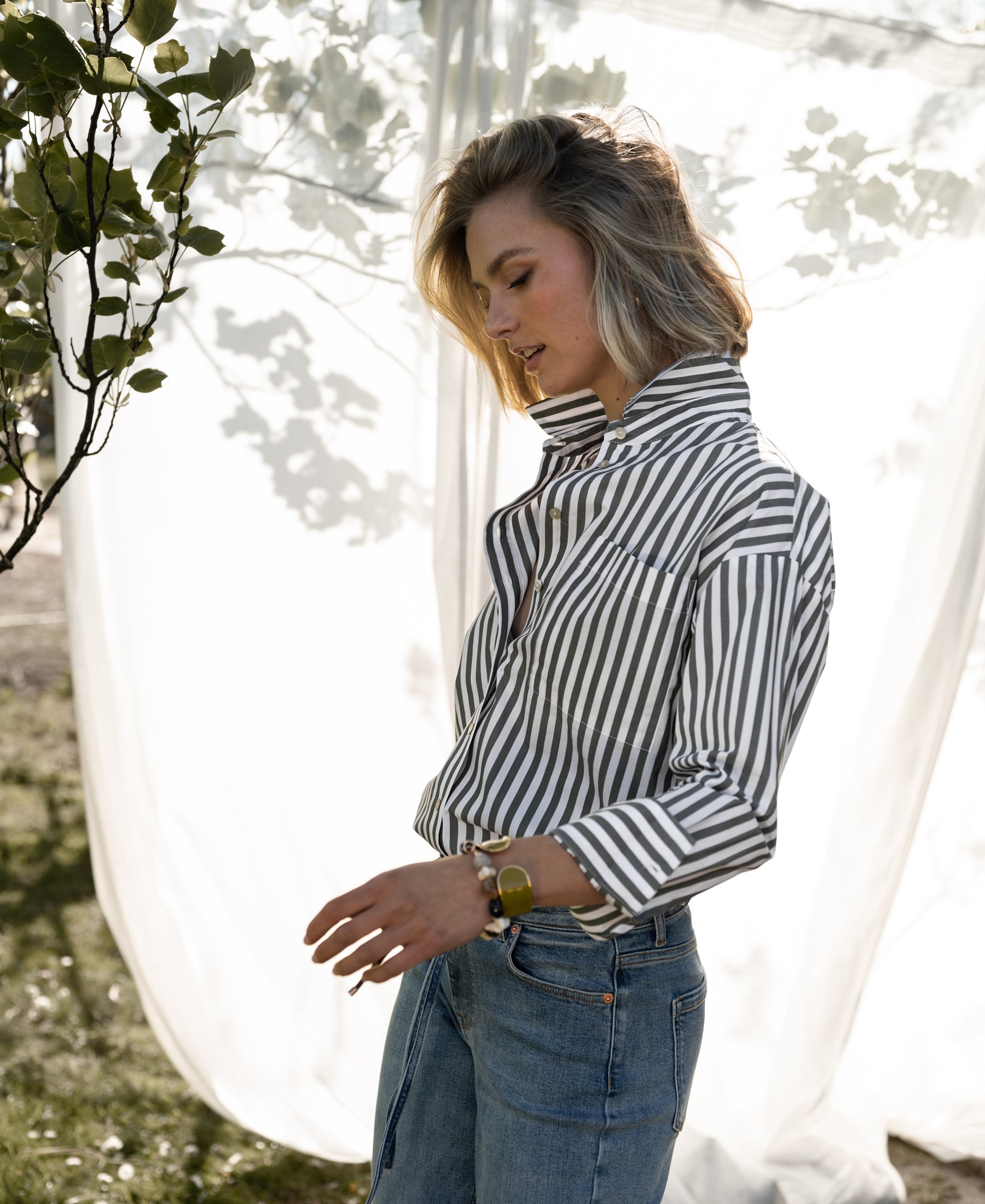 Woman wearing a PLEIN PUBLIQUE LA PASCALLE white-olive green striped blouse, standing outdoors with sunlight behind her.