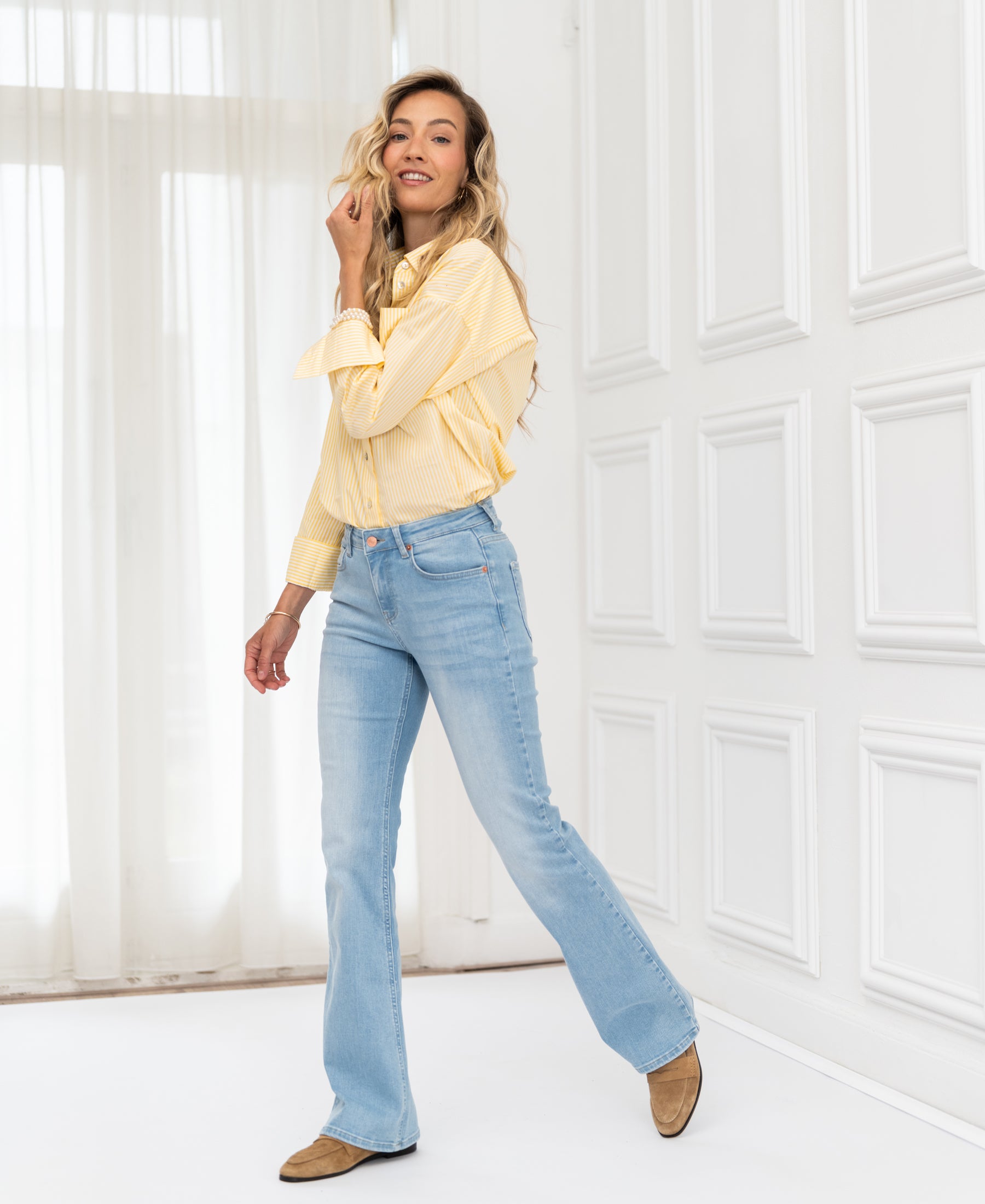 Woman wearing light-wash flared jeans indoors, smiling and touching her hair.