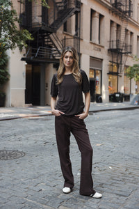 Woman wearing chocolate brown ribbed T-shirt, hands in pockets, standing on cobblestone street.