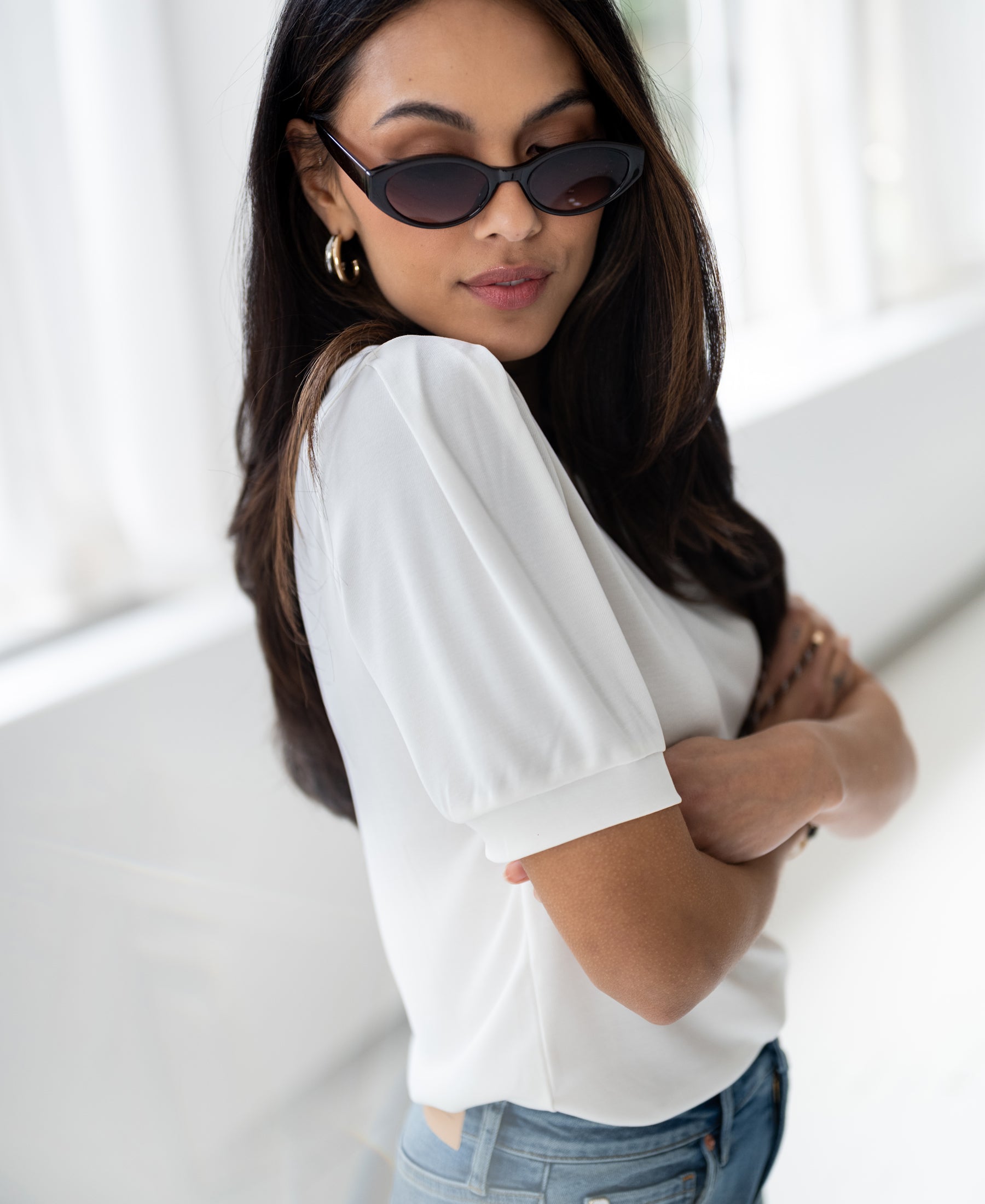 Woman wearing an ivory ribbed T-shirt stands with arms crossed in a bright room.