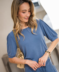 Woman wearing a ribbed blue t-shirt stands with hand on waist, smiling.