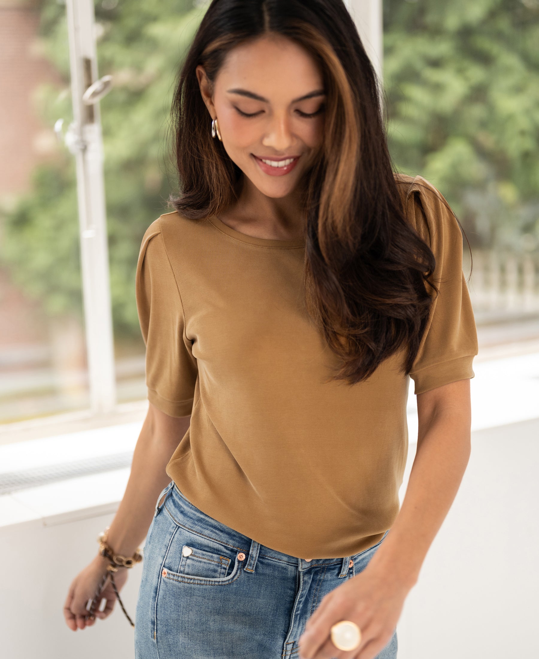 Woman wearing a yellow ribbed T-shirt called LA PHILEINE by PLEIN PUBLIQUE stands near a bright window with greenery outside.