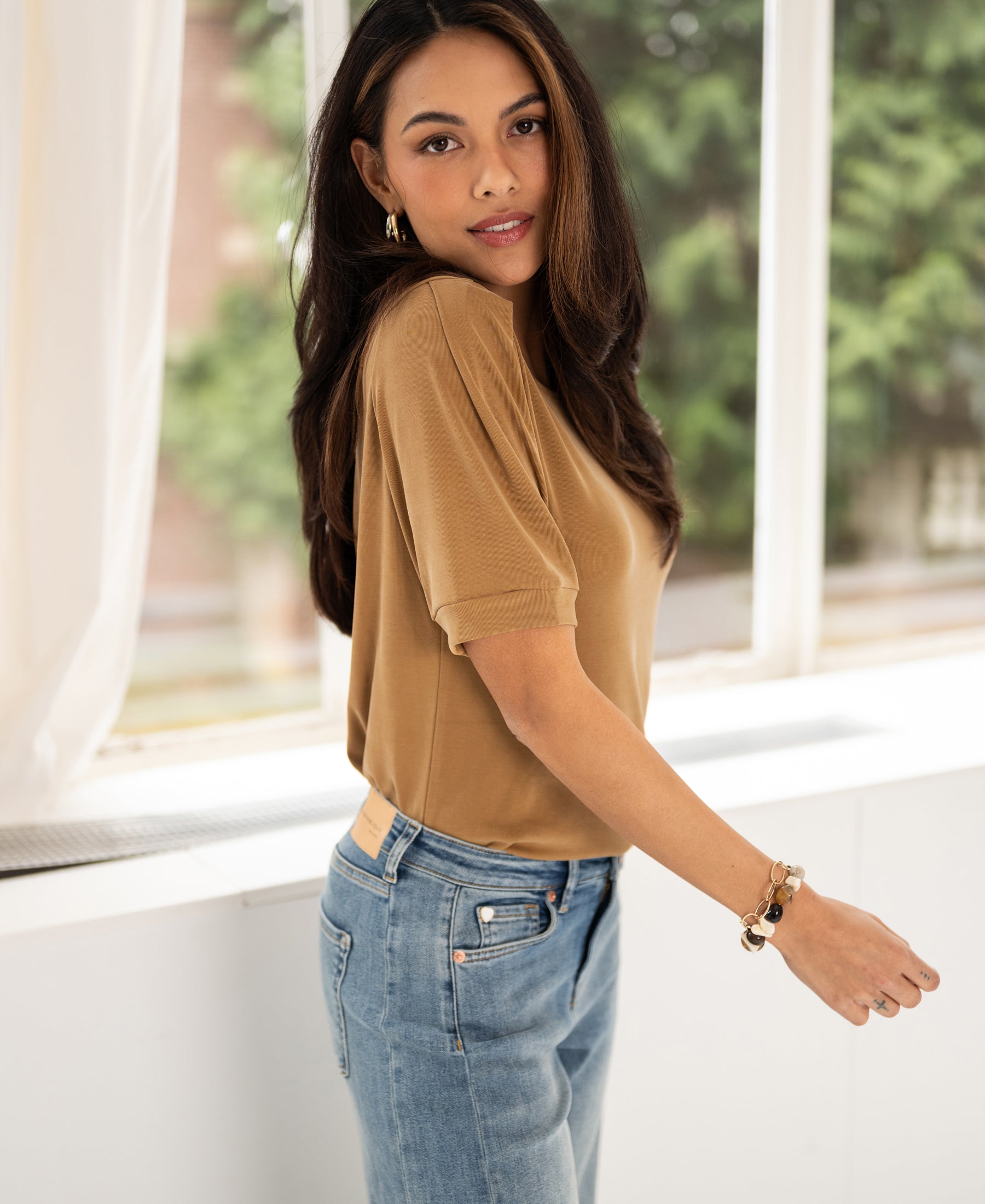Woman wears a golden yellow ribbed LA PHILEINE T-shirt by PLEIN PUBLIQUE, standing by a bright window.