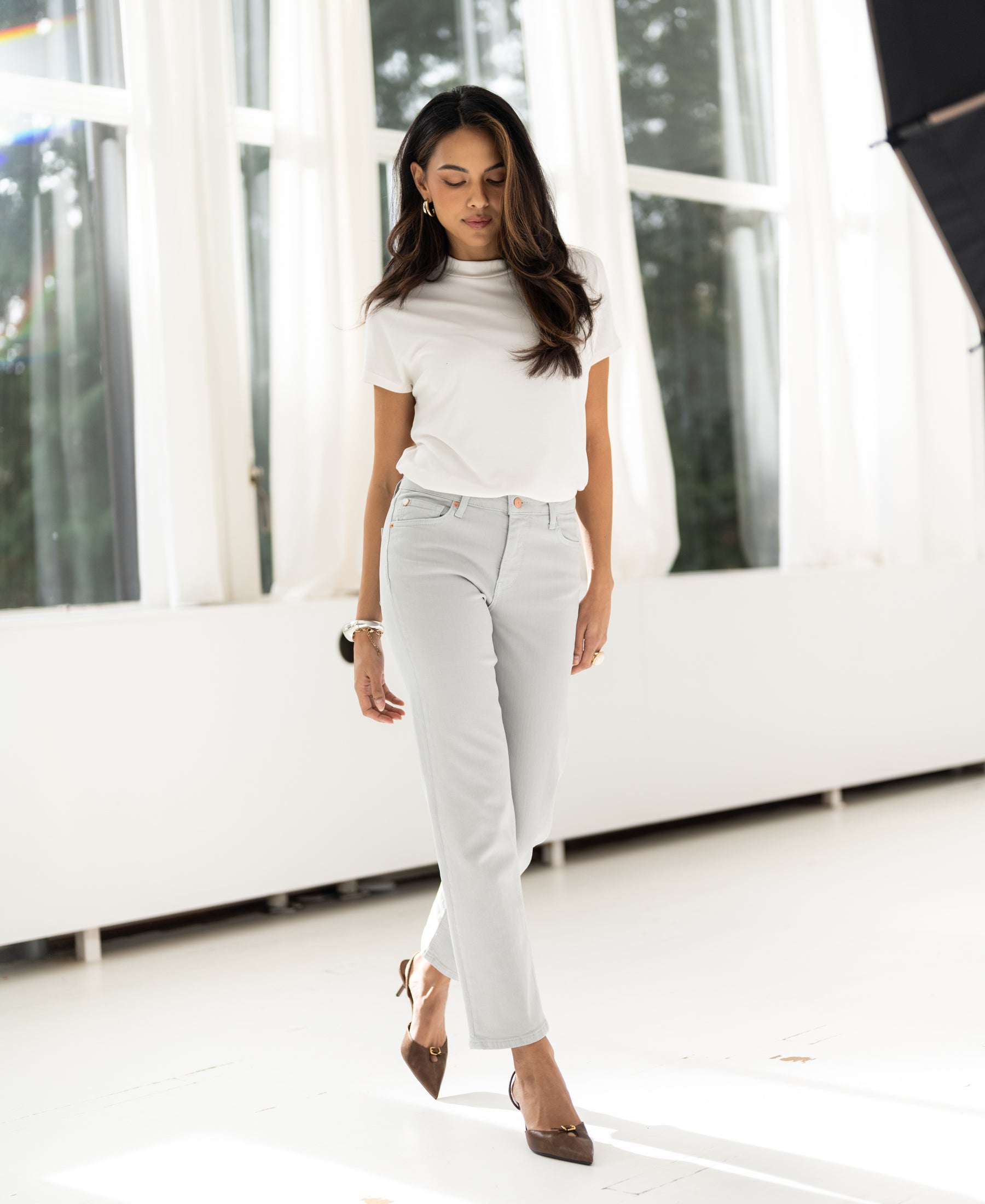 Woman wearing ivory ribbed collared top by PLEIN PUBLIQUE stands in a bright room, looking down.