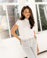 Woman wearing ivory ribbed top by PLEIN PUBLIQUE stands by windows, smiling with eyes closed.