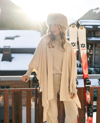 Woman wearing a light sand merino wool poncho stands outdoors on a sunny, snowy balcony.