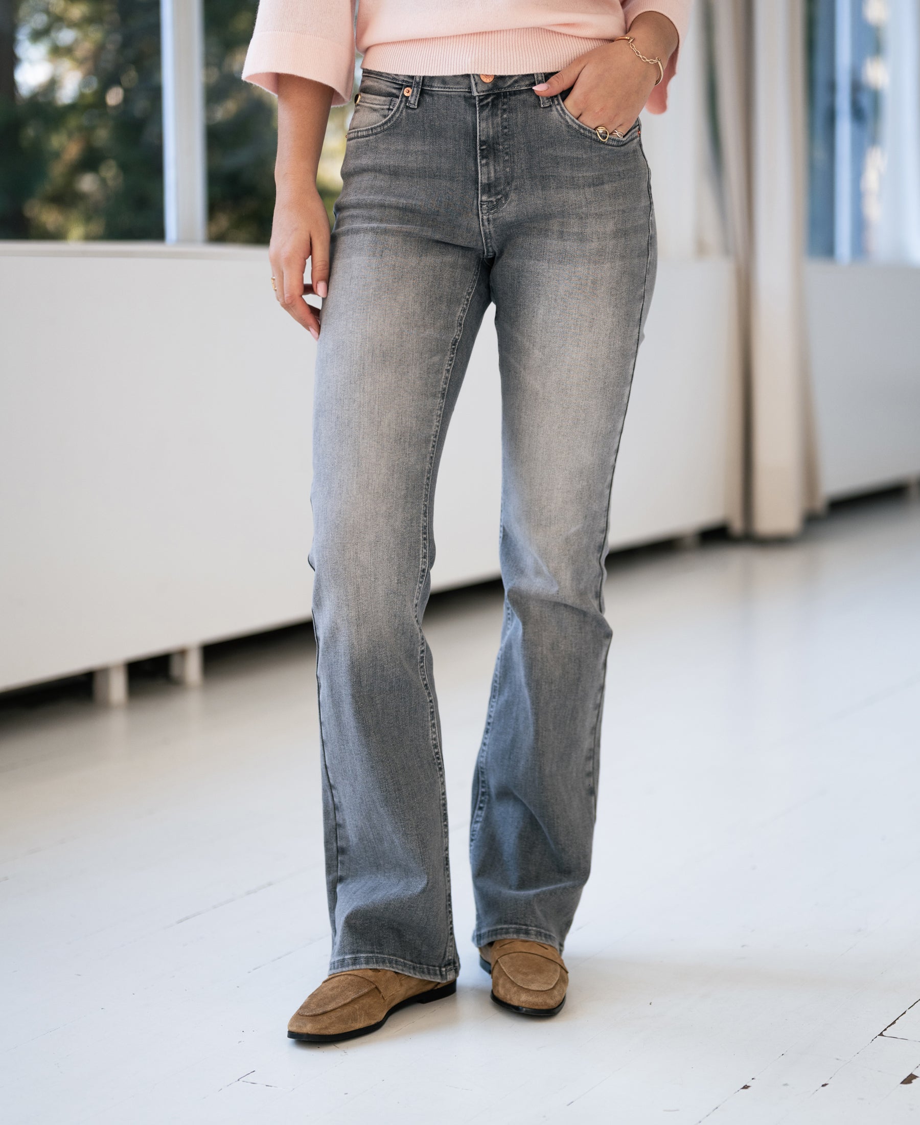 A person wearing grey stonewash flared jeans stands by large windows.