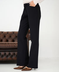 Model wears black flared pants by PLEIN PUBLIQUE, standing near a brown leather sofa.