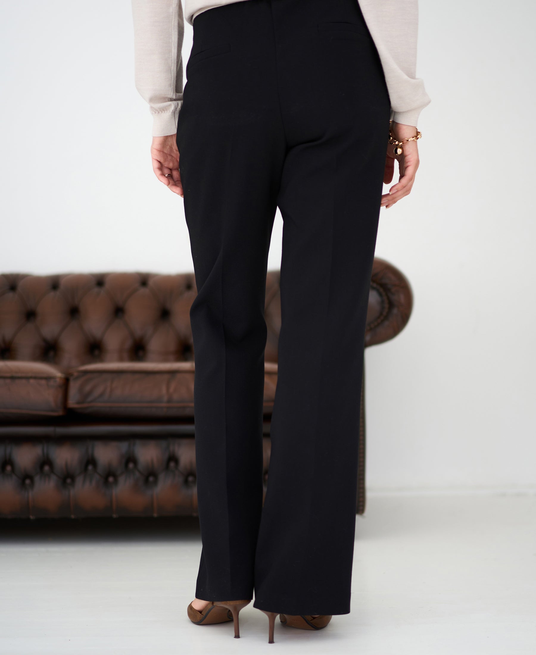 Woman stands before a brown leather sofa, wearing black flared pants.