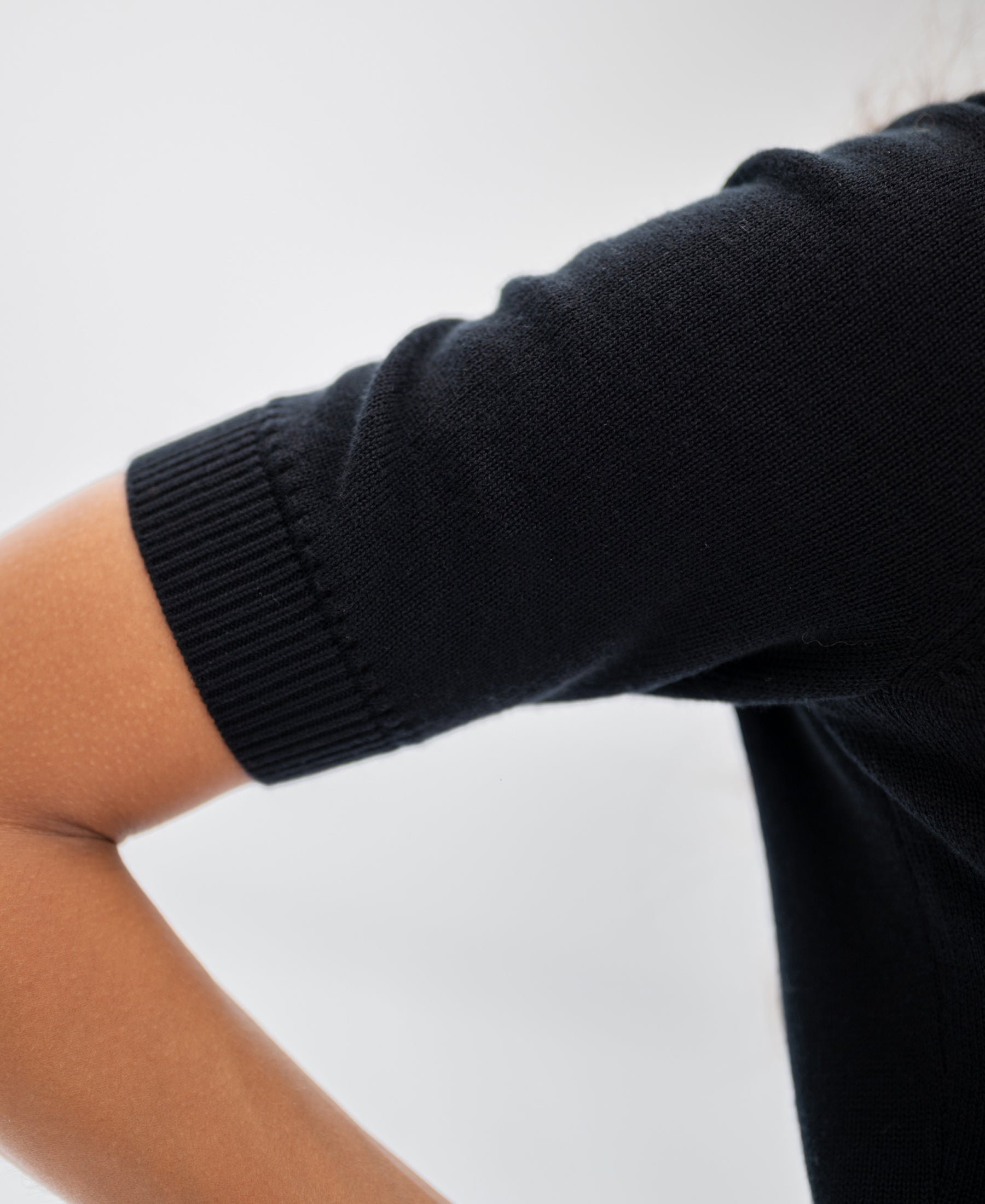 A bent arm in a black cotton-merino wool knit sweater with short sleeves on a light background.