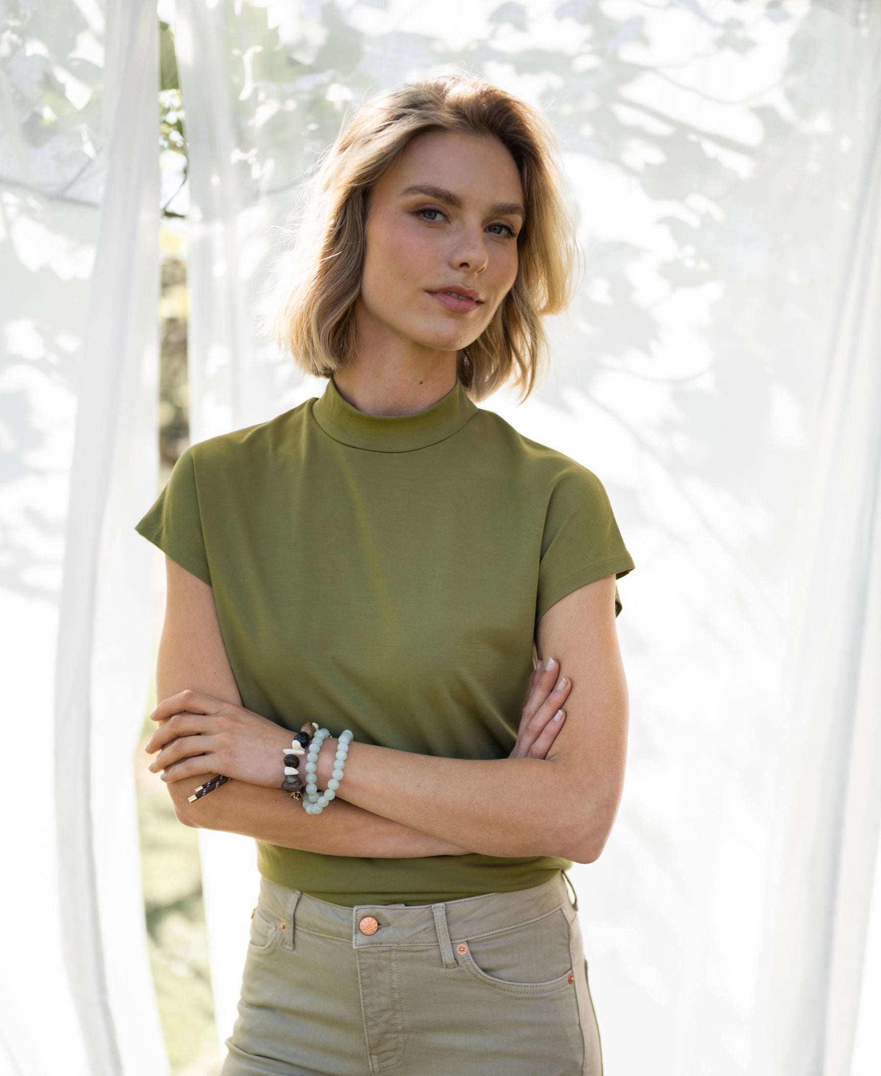 Woman wearing olive green PLEIN PUBLIQUE travel collared top, standing in front of white outdoor curtains.