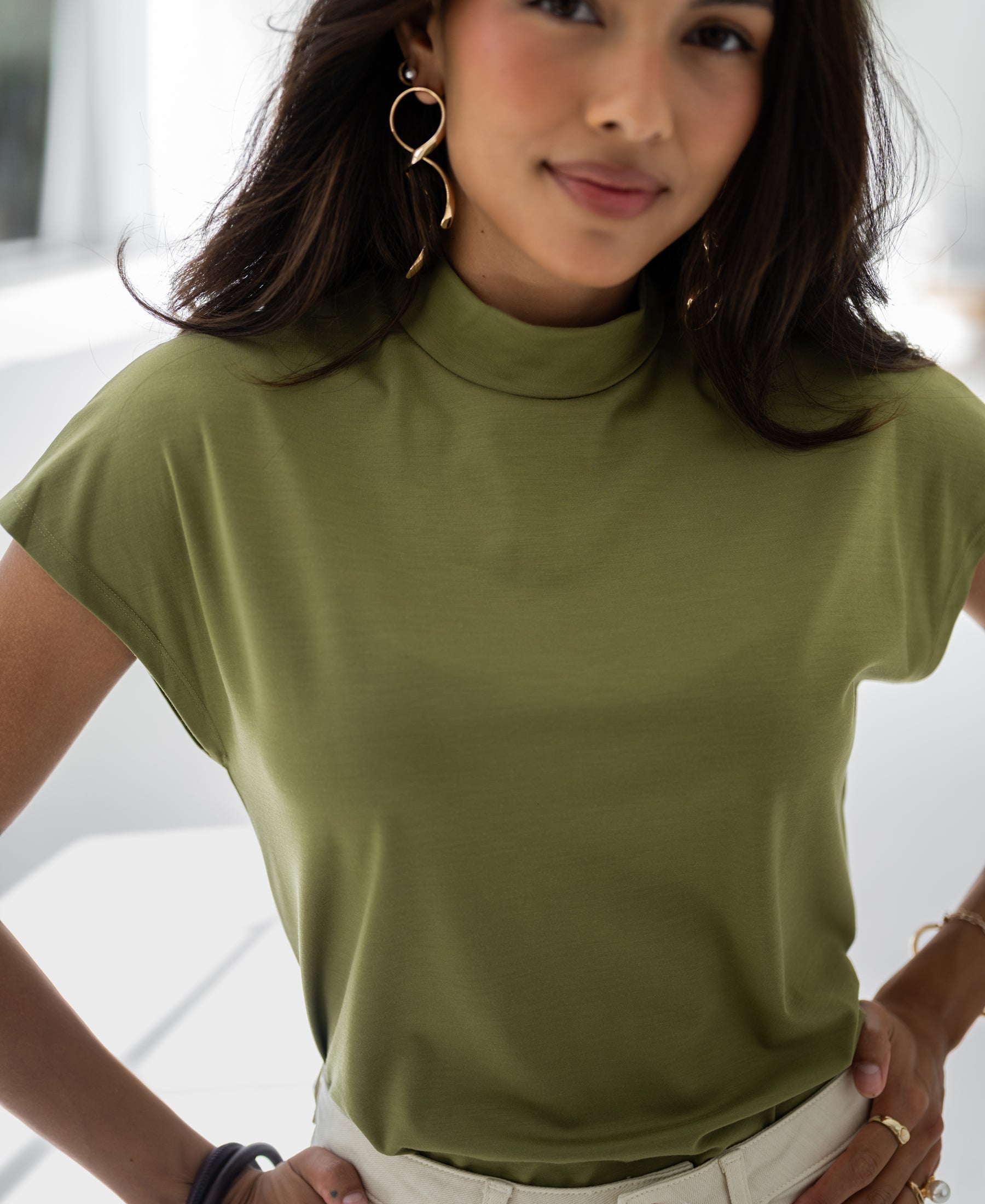Woman wearing an olive green button-up blouse, standing indoors with hands on hips.