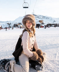 Woman wears blush PLEIN PUBLIQUE Merino turtleneck vest, kneeling on snowy ground at a ski resort.