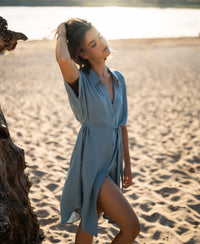 Cupro dress in denim cloud color shown outdoors with model standing on a sunny beach, eyes closed.
