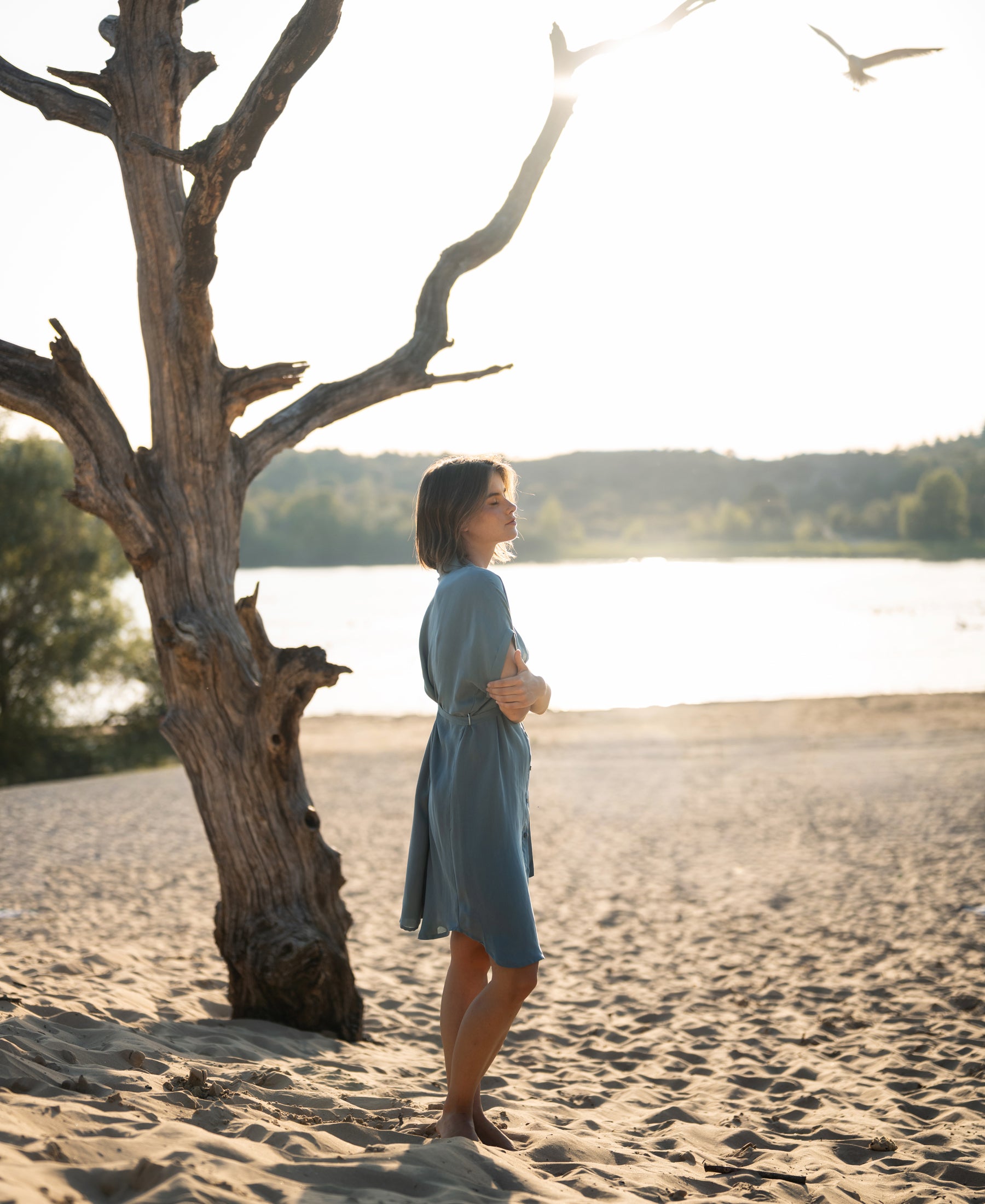 Cupro dress in light blue denim shade, shown on a beach with sunlight glimmering on the water.