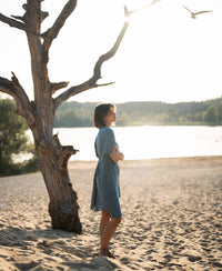 Cupro dress in denim cloud color shown on sandy beach with a tree and water in the background.