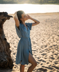Cupro dress LA SELENE in Denim Cloud color shown on a woman standing on a sandy beach near a tree trunk.