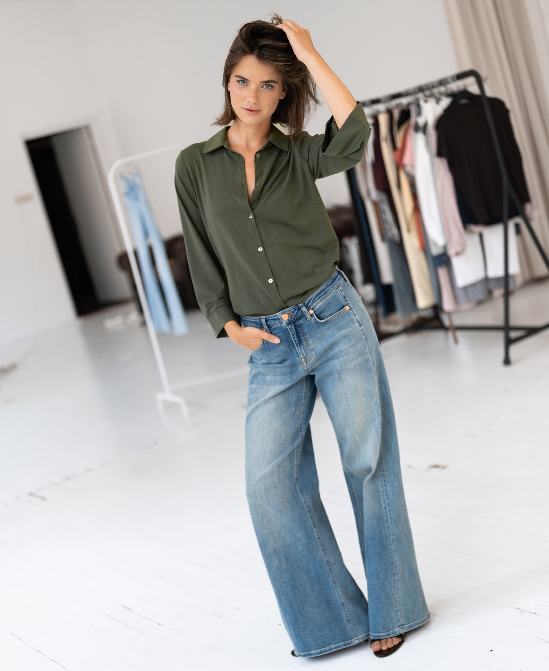 Woman wearing olive green Travel Blouse LA SILVESE by PLEIN PUBLIQUE stands in a bright room with racks in the background.