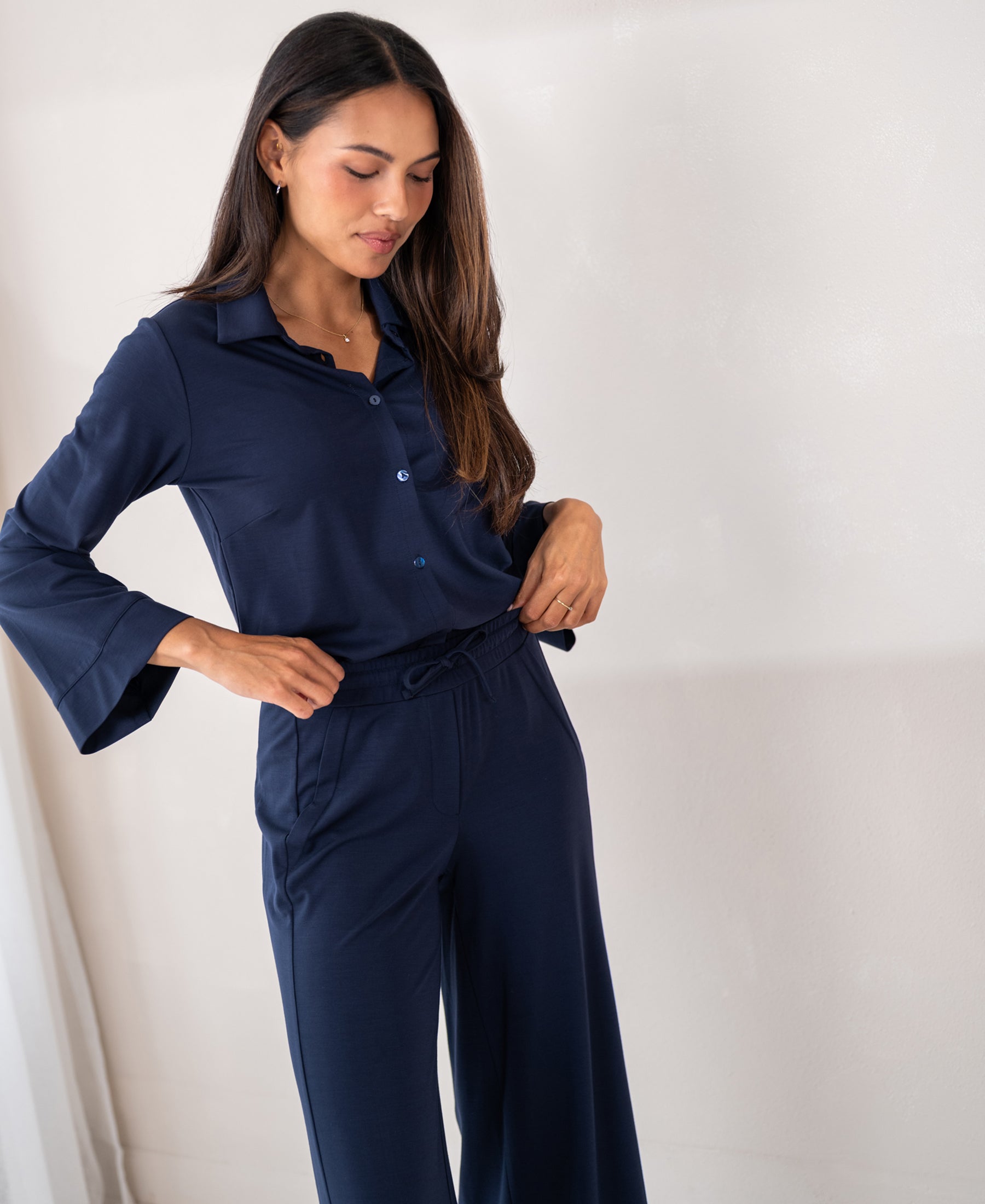 Woman wearing navy PLEIN PUBLIQUE LA SILVESE travel blouse, standing by a light wall.