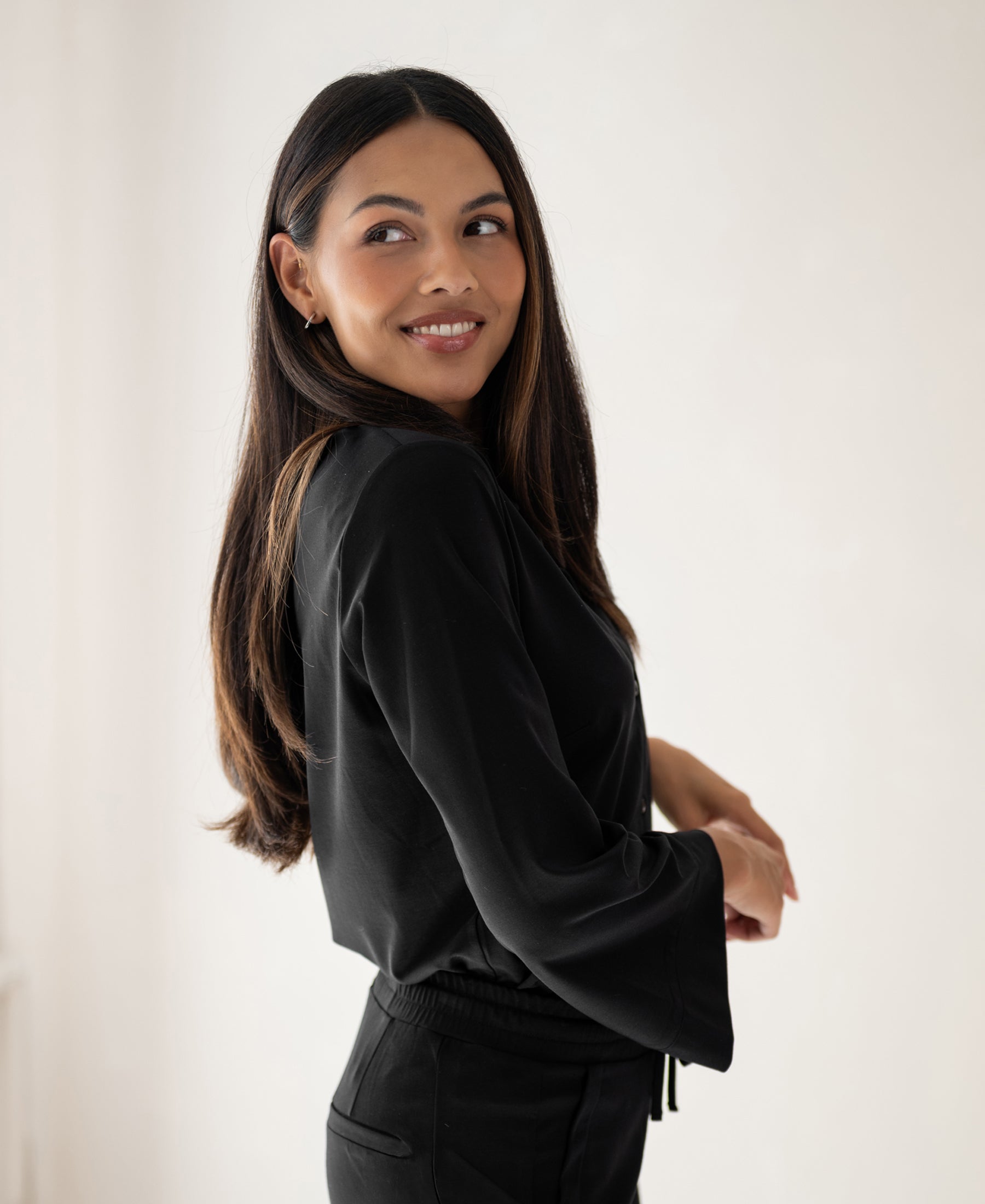 Woman wearing a black PLEIN PUBLIQUE travel blouse, smiling over her shoulder.