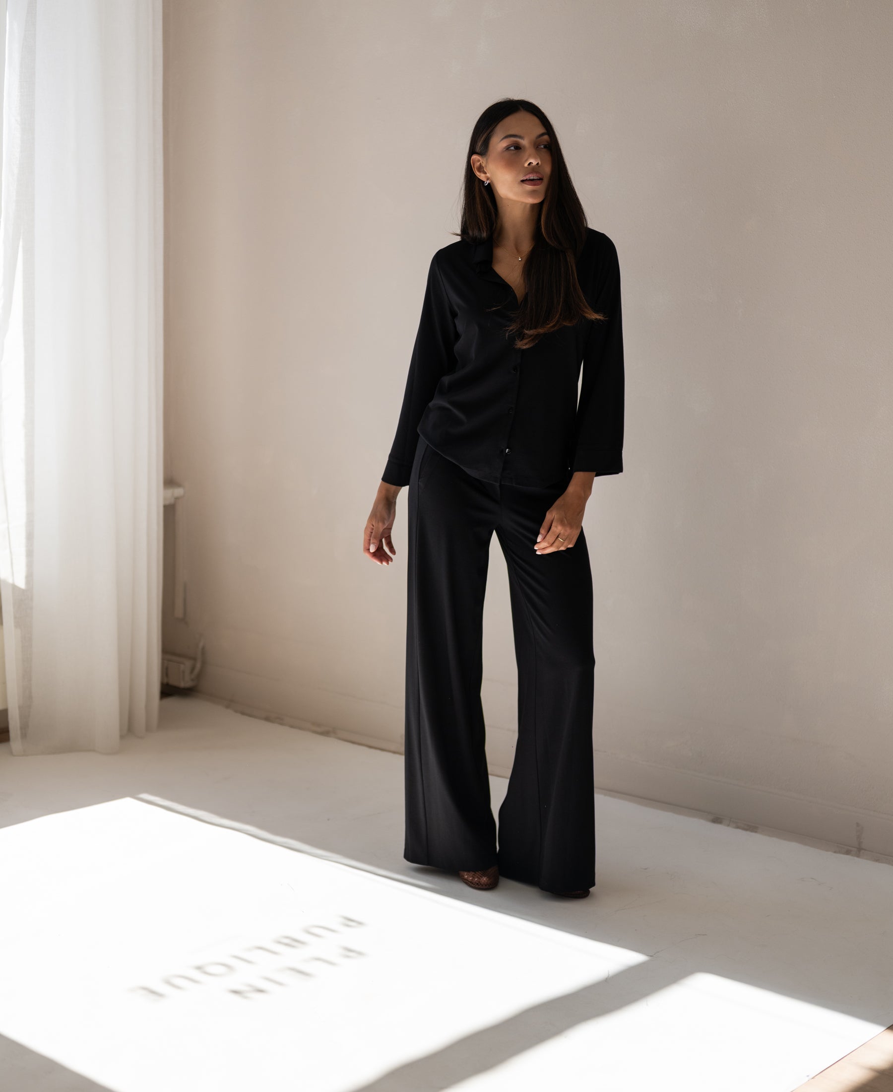 Black travel pants LA JULIA by PLEIN PUBLIQUE, shown indoors with sunlight streaming through a window.