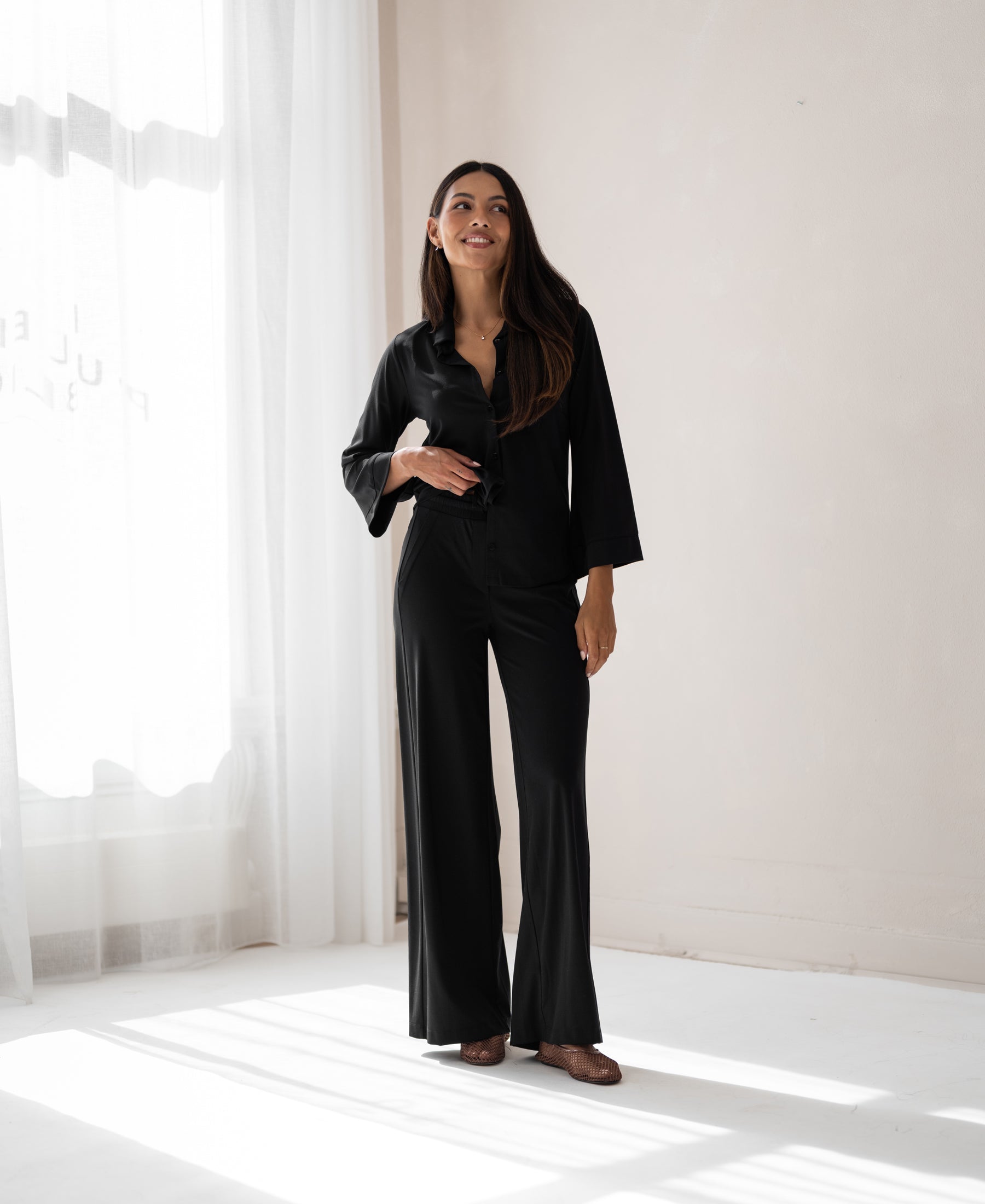 Black PLEIN PUBLIQUE LA JULIA travel pants displayed in a bright, minimalist room.