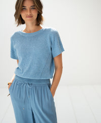 Woman wears the linen summer sweater LE SOLEIL in light blue, smiling in a minimal space.