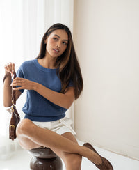 Woman wearing linen summer sweater THE SUN by PLEIN PUBLIQUE, sitting relaxed on a stool.