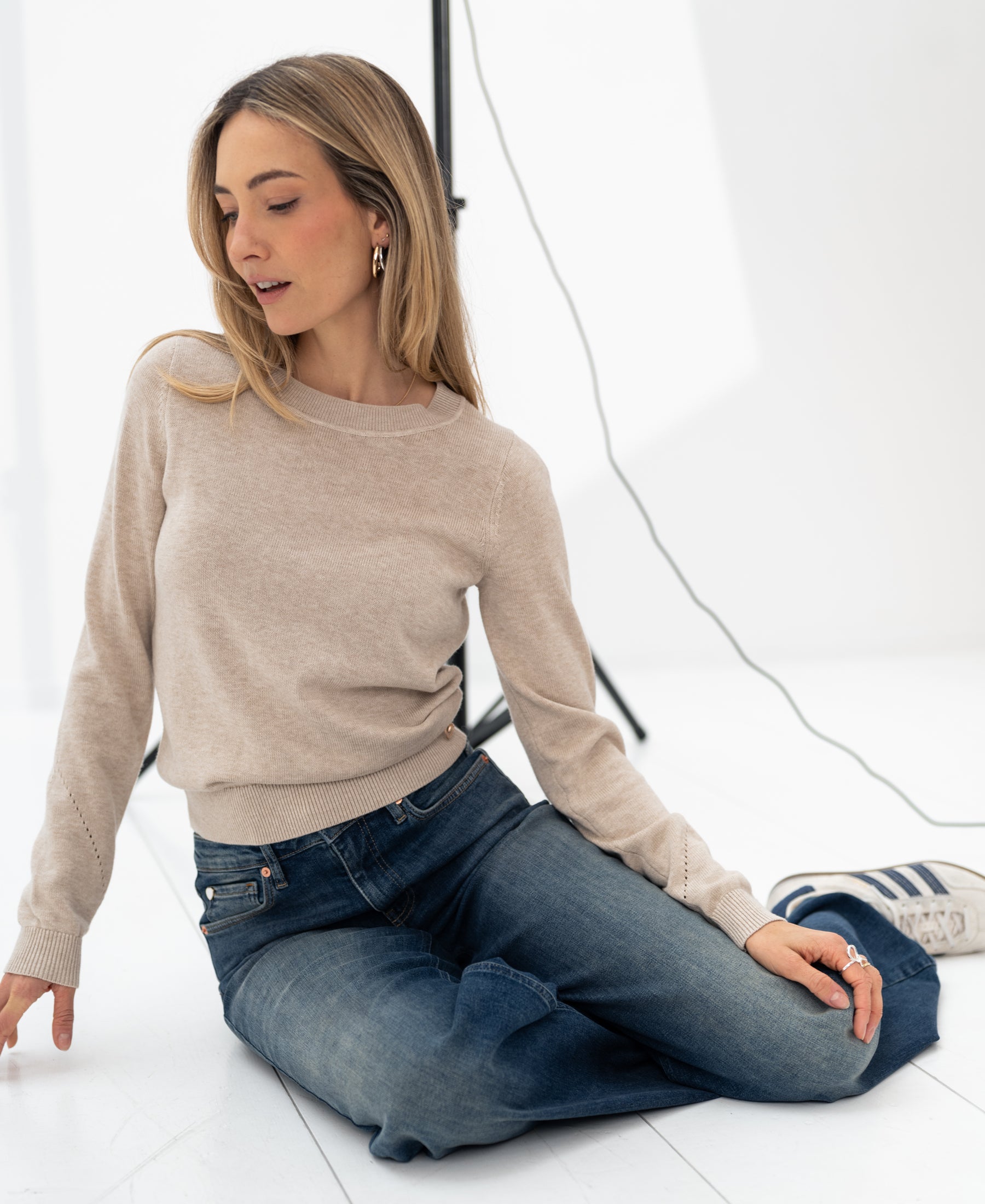 Woman wearing a beige merino wool sweater sits on a white floor, studio lights behind her.