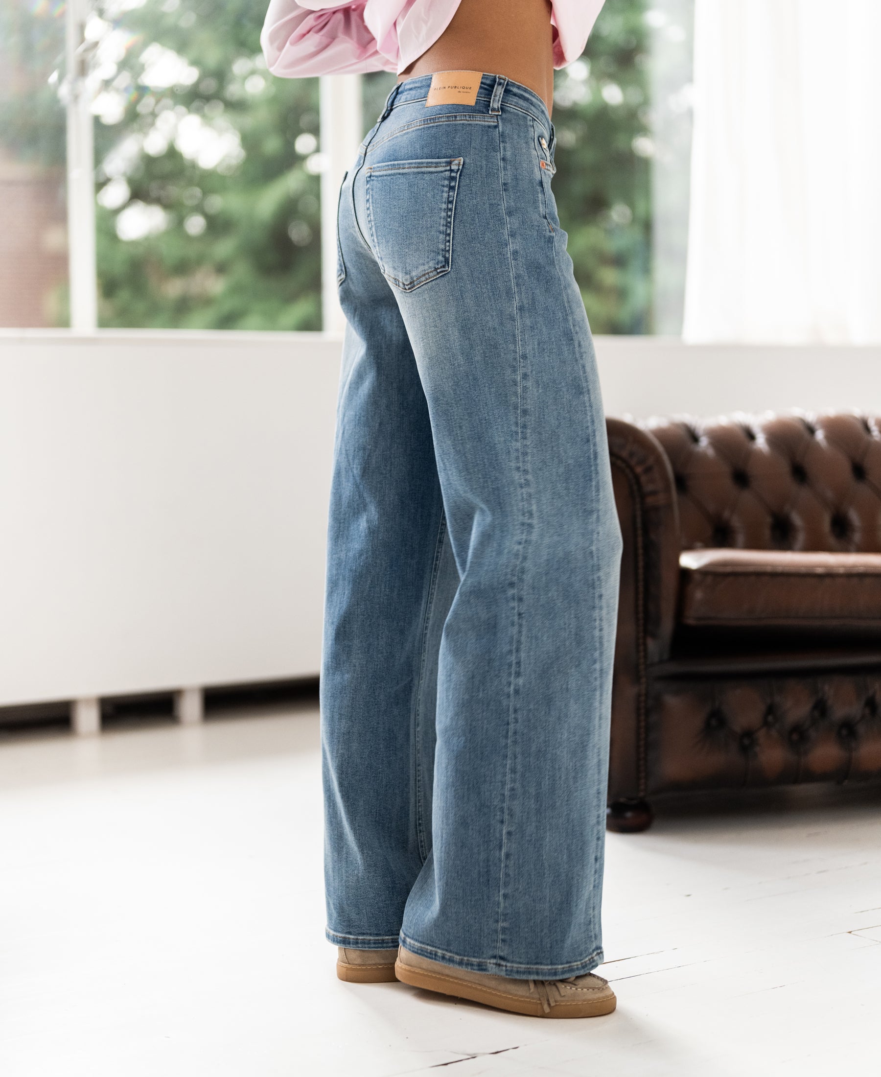 Wide leg jeans in light stone color shown in a bright room with a brown couch.