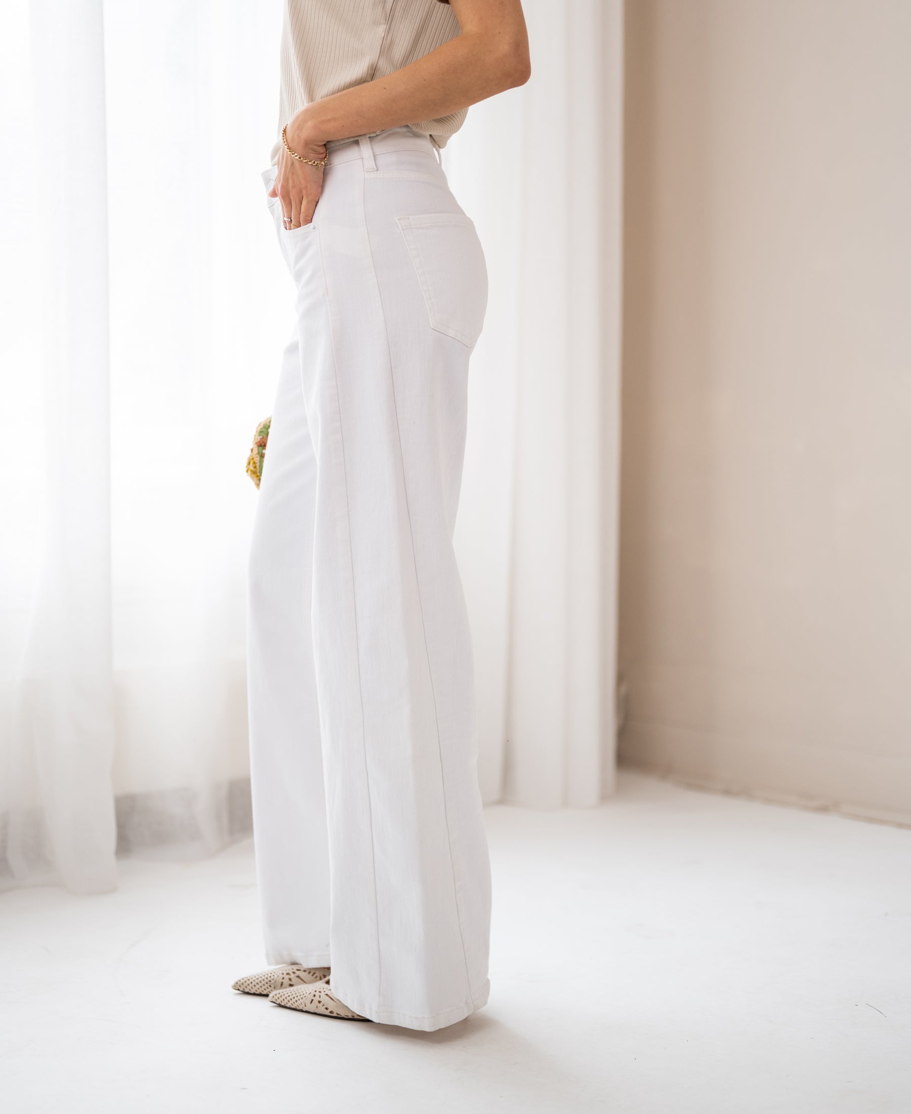 Woman wearing wide leg white jeans stands indoors by a white curtain.