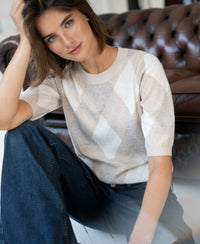 Woman wearing sand melange argyle merino wool sweater, smiling in front of a brown leather sofa.