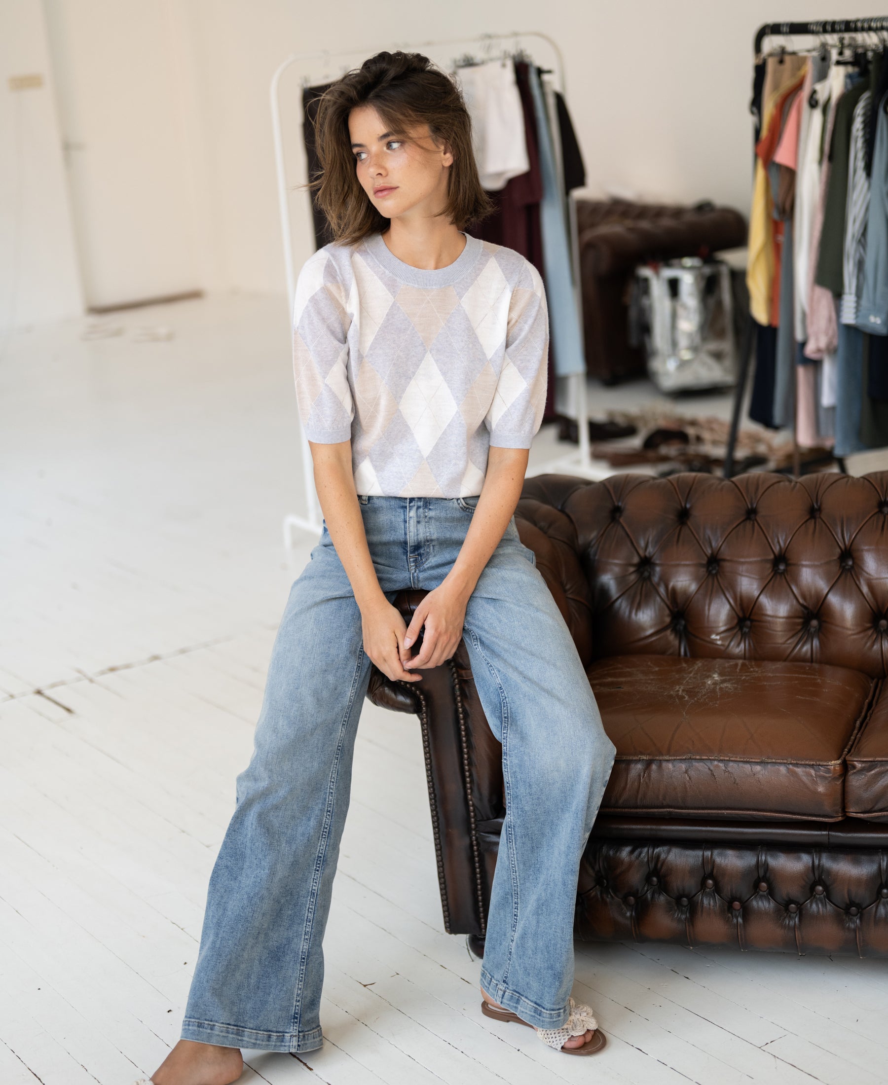 Woman wearing PLEIN PUBLIQUEs light grey argyle merino wool sweater sits on a leather couch.